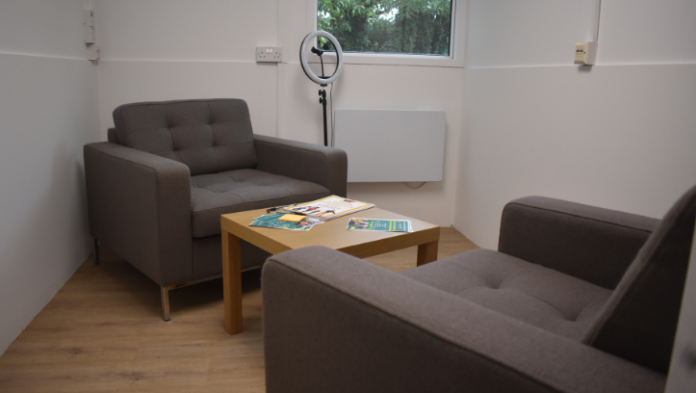 A small, quiet meeting room with two armchairs facing each other, a table with leaflets, and a ring light near a window, suggesting a space for private conversations or consultations.