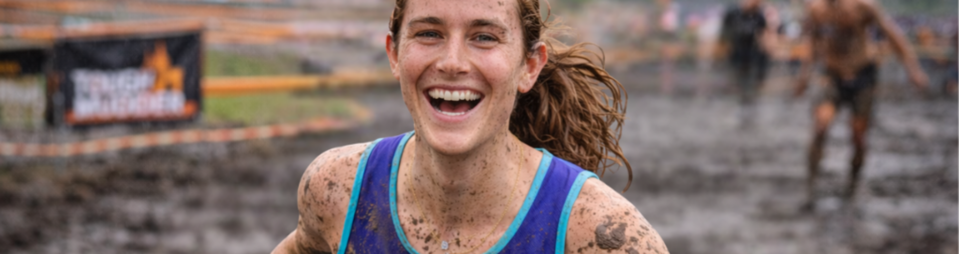 A woman laughs while running through a muddy obstacle course at a Tough Mudder event, covered in mud and wearing a blue running vest, with other participants visible behind her