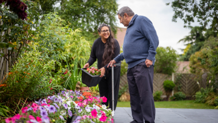 A younger woman smiles warmly while holding a tray of plants alongside an older man using a walking stick, as they tend to a colourful flower border together in a garden.