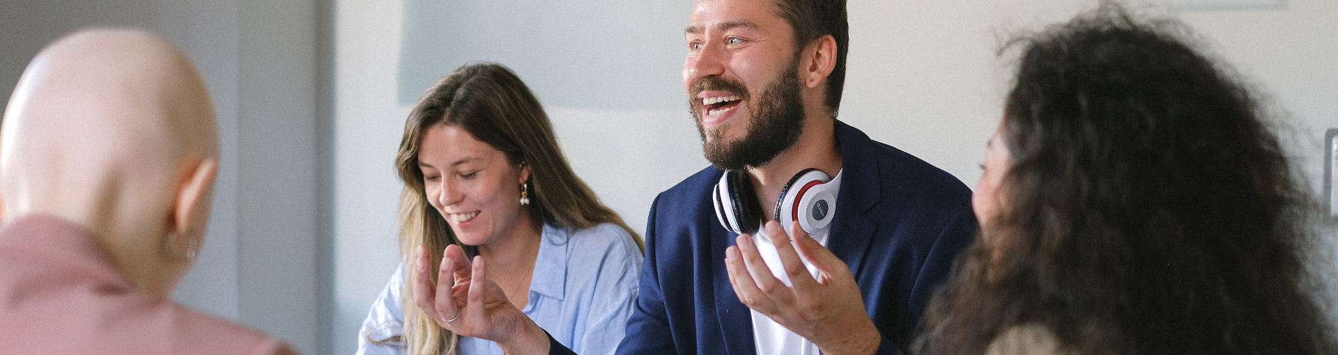 A group of colleagues laugh and engage animatedly in a bright meeting room, with one man wearing headphones around his neck.