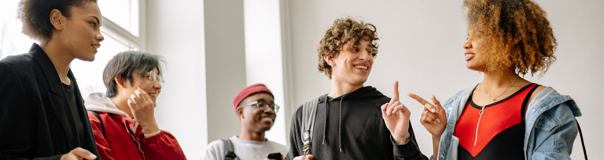 A diverse group of young people standing together indoors, smiling and talking, with two people gesturing playfully while others listen and laugh.
