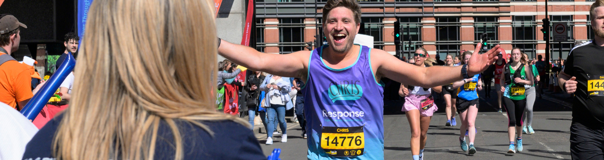 A Response runner named Chris spreads his arms wide and beams with joy during a road race, reaching out towards a supporter on the sideline, with other runners and crowds visible behind him.