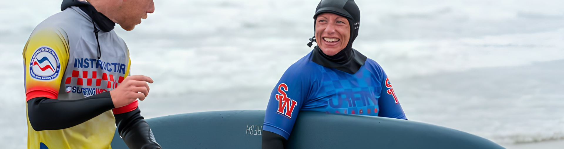 A woman in a wetsuit laughs with a surf instructor on the beach, holding a surfboard, with the sea behind them