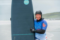 A woman in a wetsuit and neoprene hood smiles warmly while hugging a surfboard on a beach, with the sea visible behind her.