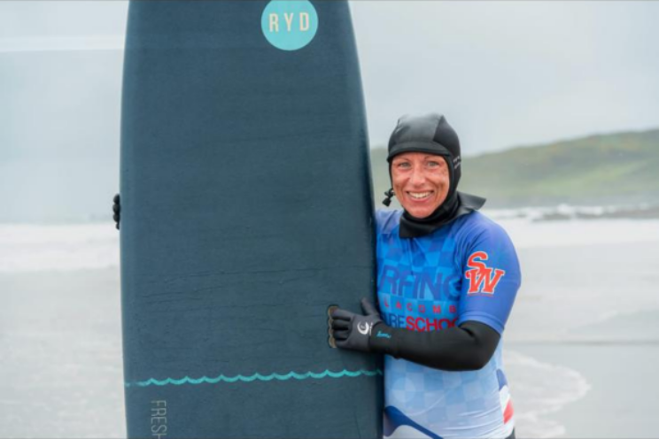 A woman in a wetsuit and neoprene hood smiles warmly while hugging a surfboard on a beach, with the sea visible behind her.