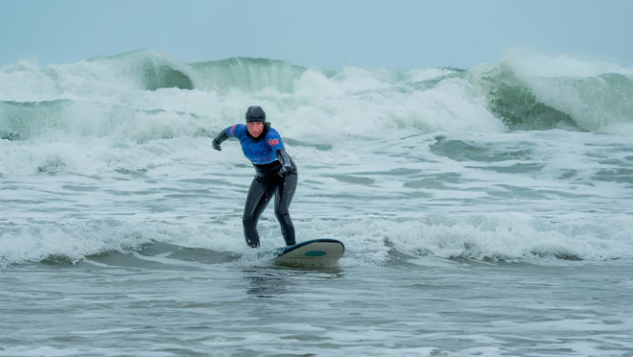 A woman in a wetsuit rides a wave on a surfboard in choppy grey seas, with large waves breaking behind her.