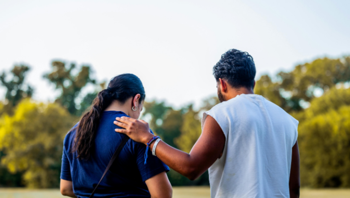 Two people seen from behind in an open green space, one resting a supportive hand on the other's shoulder in a warm evening light.