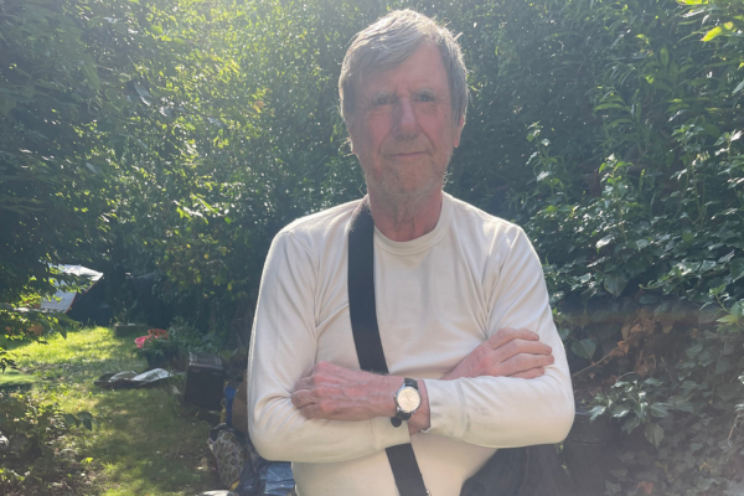 An older man stands with his arms folded in a sunlit garden, looking directly at the camera with a calm, composed expression.