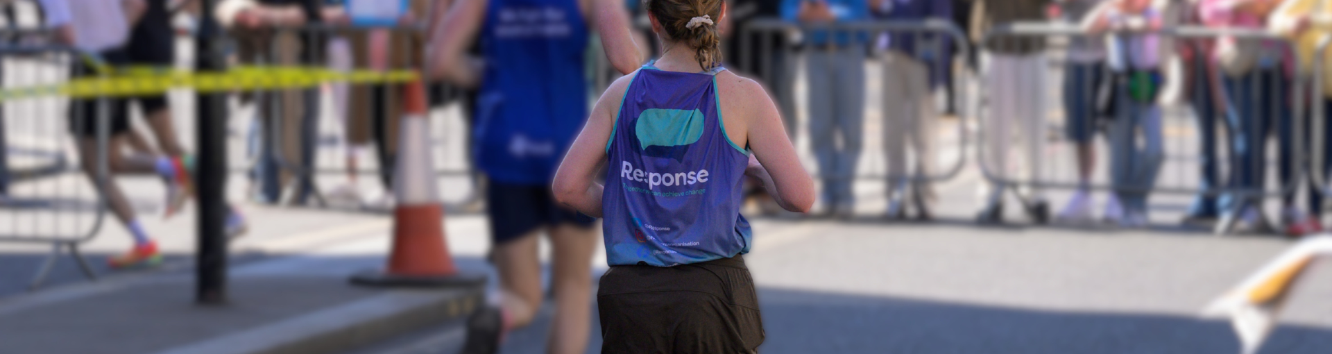 A Response runner seen from behind, wearing a purple and teal Response vest, making their way along a road race route lined with spectators.