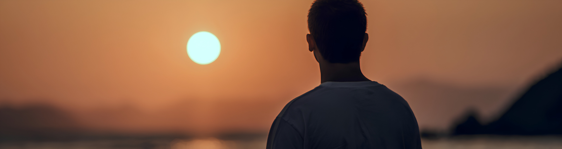 A person seen from behind, silhouetted against a warm golden sunset over water, standing quietly in a moment of reflection.