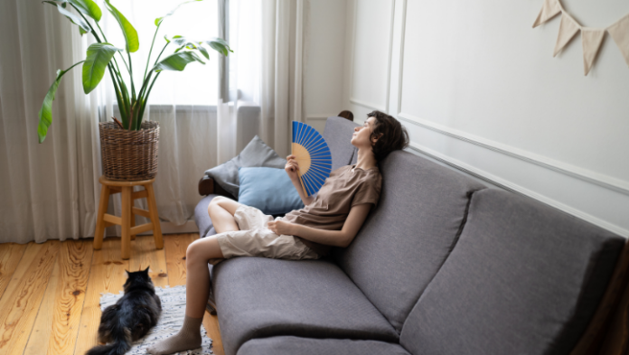 A young woman reclines on a sofa fanning herself with a handheld fan on a hot day, with a cat resting on the floor beside her in a bright, plant-filled living room.