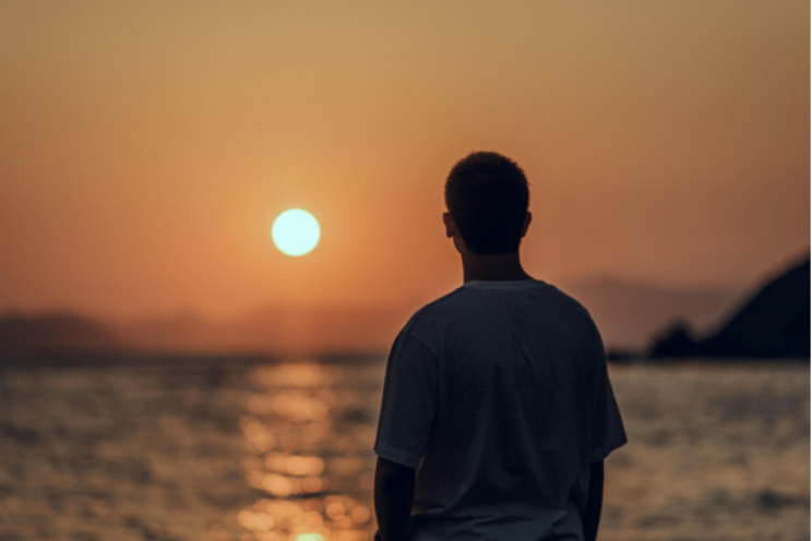 A person seen from behind, silhouetted against a warm golden sunset over water, standing quietly in a moment of reflection.