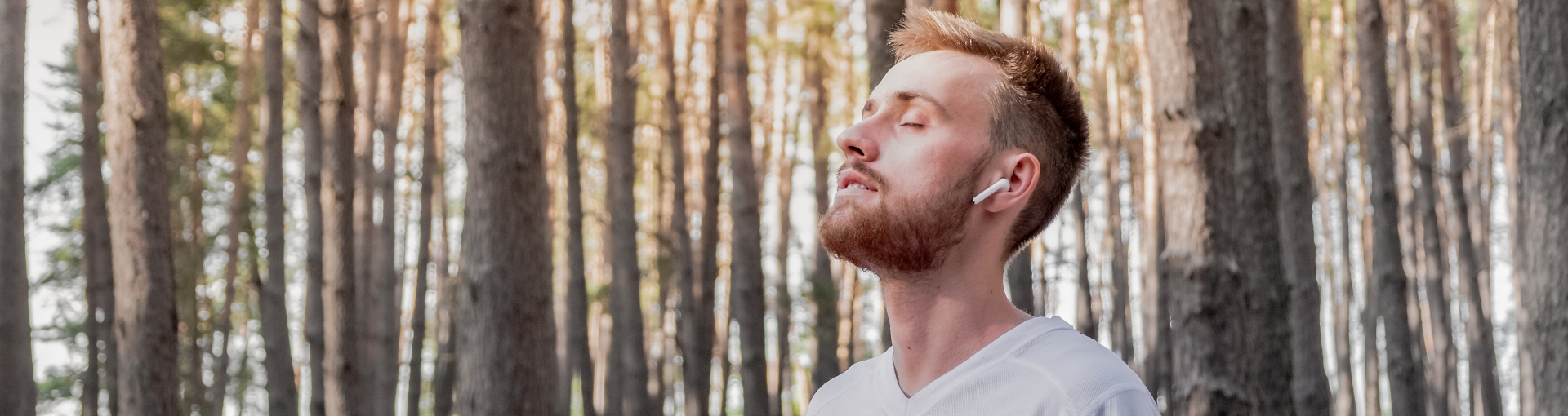 A young man stands with his eyes closed and face tilted upward among tall pine trees, wearing earbuds and a white t-shirt in a moment of calm and stillness.