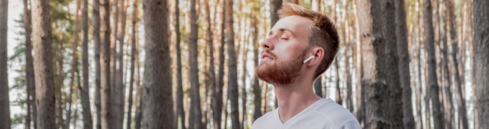 A young man stands with his eyes closed and face tilted upward among tall pine trees, wearing earbuds and a white t-shirt in a moment of calm and stillness.