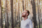 A young man stands with his eyes closed and face tilted upward among tall pine trees, wearing earbuds and a white t-shirt in a moment of calm and stillness.
